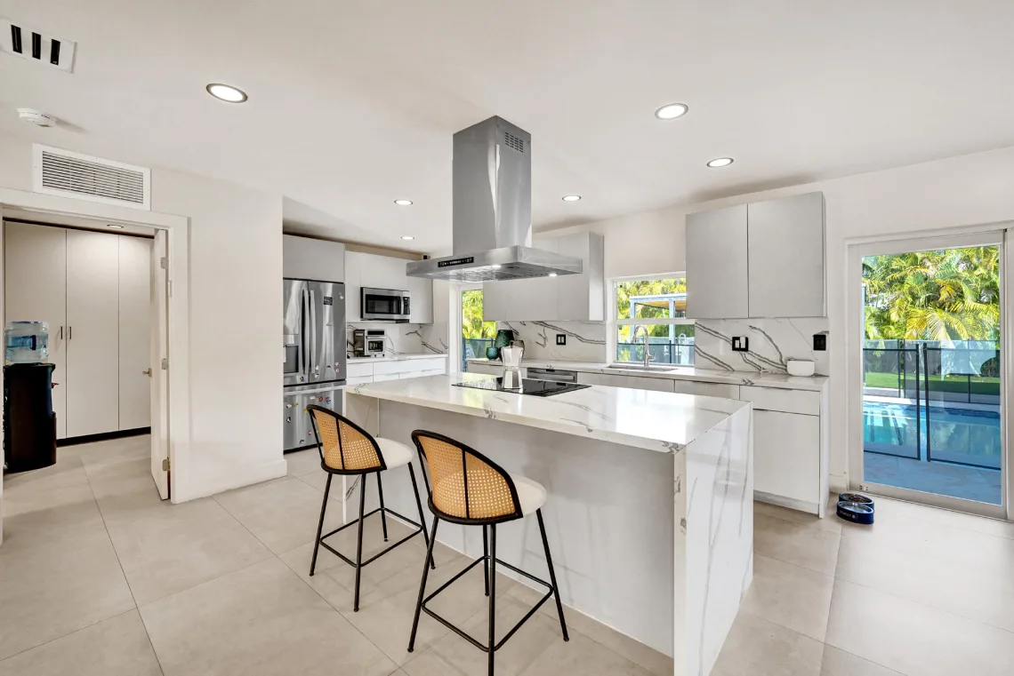 White kitchen with marble waterfall island, stainless hood, refrigerator, and pool-view door