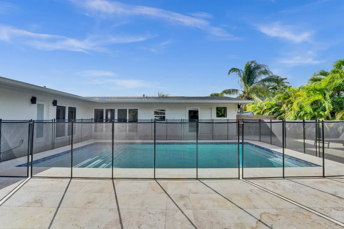 Rectangular pool and travertine deck behind a single-story white home with palms