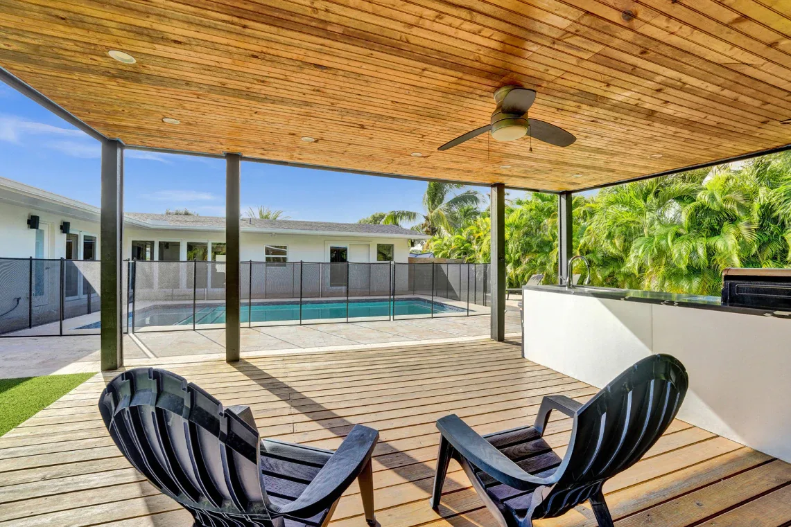 Backyard pool viewed from covered wood-clad deck with ceiling fan and lounge chairs