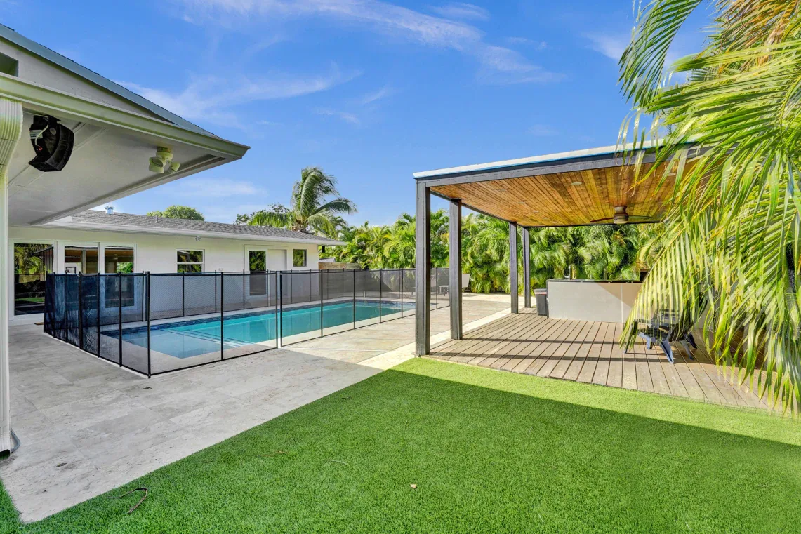 Fenced backyard pool with wood pergola, artificial turf, and tropical landscaping